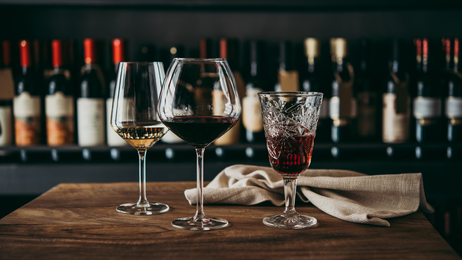 This image shows three different glasses of wine filled with white, red, and rose wine respectively. In the background a wall filled with bottles of wine.